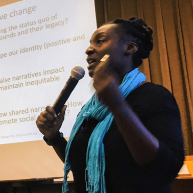Black woman with pinned up dreadlocs and blue scarf speaks before a crowd